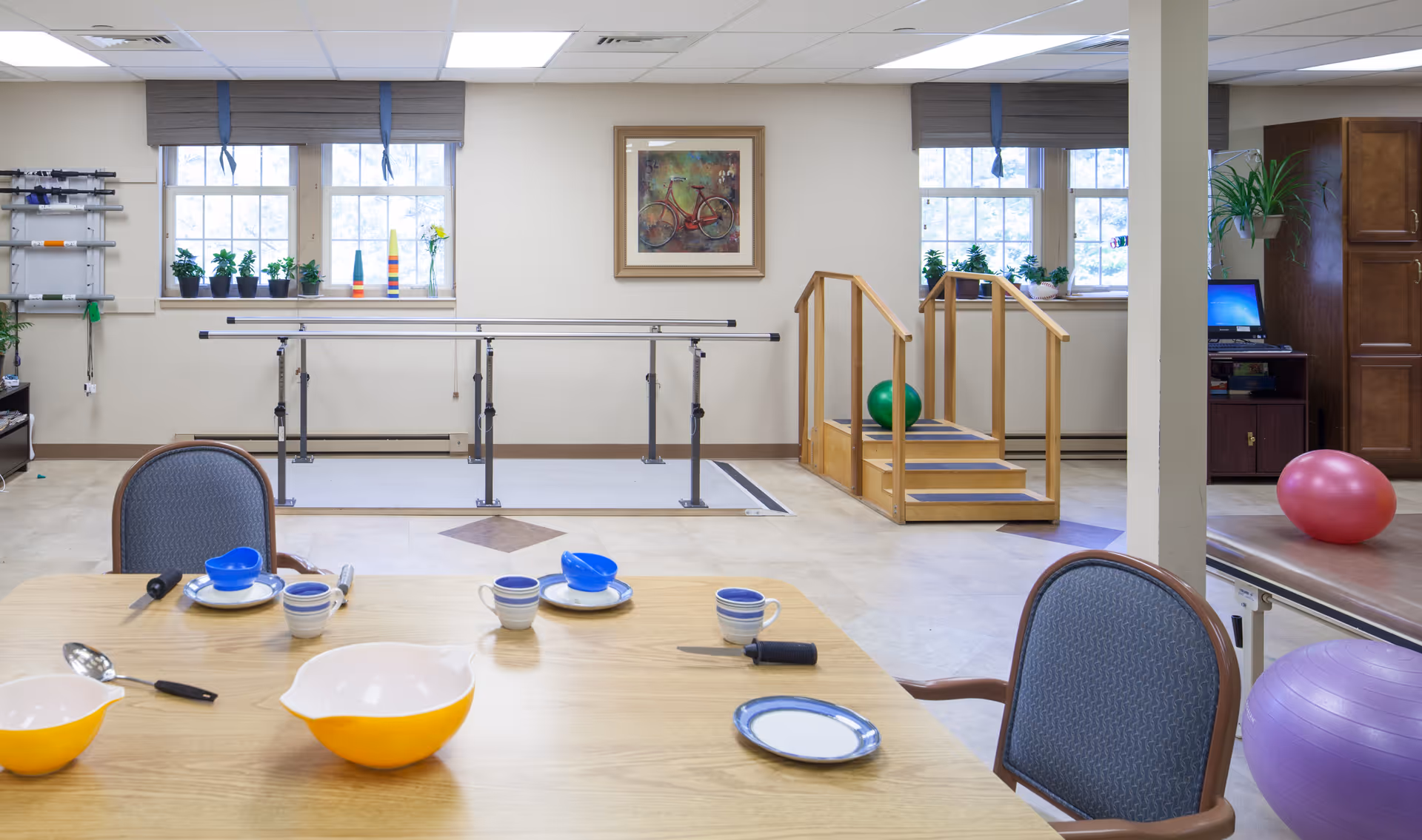 A therapy room with parallel bars and wooden stairs for physical rehabilitation, exercise balls, a table set with bowls, plates, cups, and utensils, and windows with potted plants on the sills. There is a framed picture of a bicycle on the wall and a computer workstation in the background.