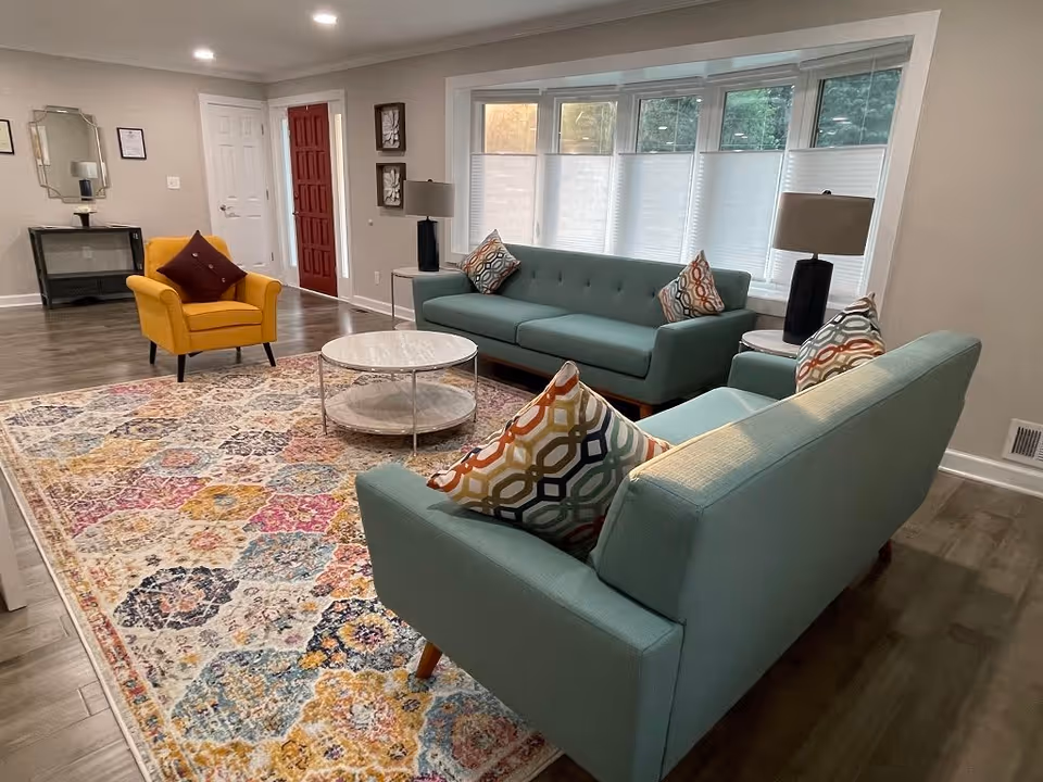 A bright and spacious living room with two teal sofas and a yellow armchair arranged around a round marble coffee table on a colorful patterned rug. The room features large windows with white blinds, two side tables with black lamps, a red front door, and a small console table with a mirror and framed certificates on the wall.