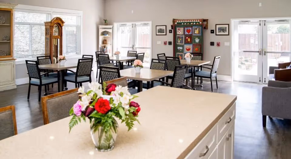 A bright and spacious dining area in a senior living facility with several tables and chairs arranged neatly. A vase with colorful flowers is placed on a countertop in the foreground. The room features large windows, a grandfather clock, a quilt hanging on the wall, and double glass doors leading outside.