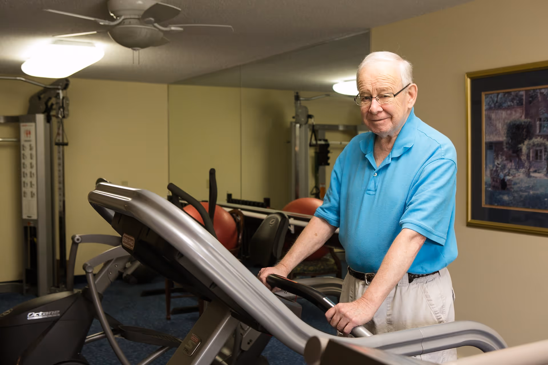 An elderly man wearing glasses and a blue polo shirt is standing on a treadmill in a fitness room. The room has exercise equipment, a ceiling fan, and a framed painting on the wall.