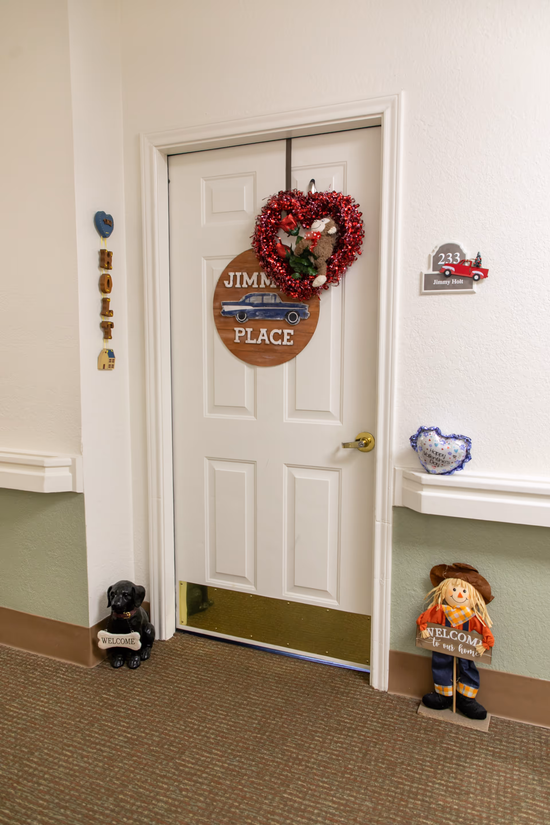 A white door in a senior living facility hallway decorated with a red heart-shaped wreath and a wooden sign that reads 'Jimmy Place'. Beside the door, there is a small black dog figurine holding a 'Welcome' sign and a scarecrow figure holding a 'Welcome to our home' sign. The door number plaque reads '233 Jimmy Holt'.
