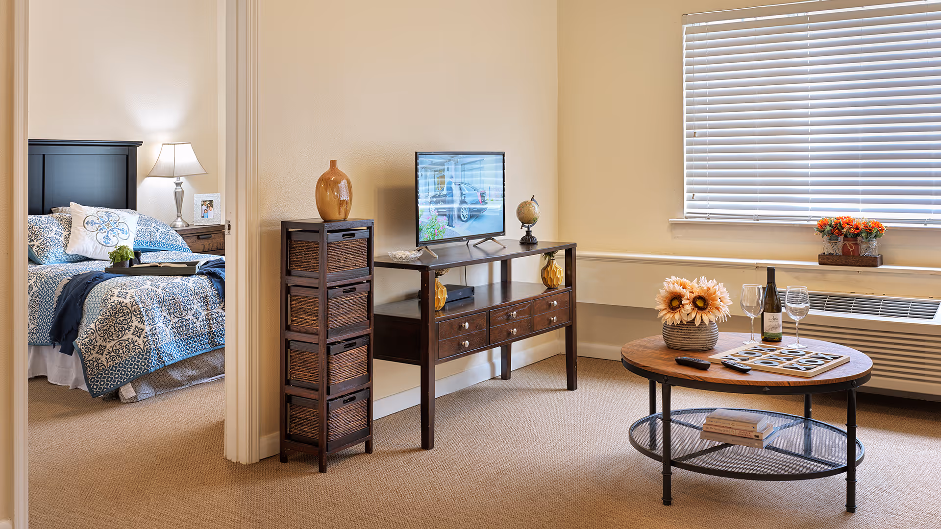 A cozy living room area with a round wooden coffee table decorated with a vase of sunflowers, a bottle of wine, and two wine glasses. A TV sits on a wooden console table with decorative items including a globe and a vase. To the left, there is a view into a bedroom with a bed covered in blue and white patterned bedding, a nightstand with a lamp, and a framed photo. The room has beige walls and carpet, and a window with closed blinds.