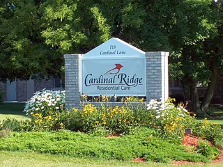 Entrance sign reading "Cardinal Ridge Residential Care" set among flower beds with trees and lawn in front of the building.
