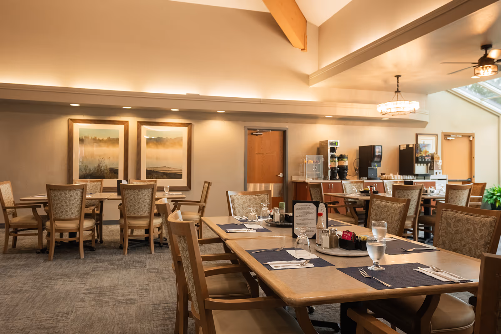 A well-lit dining room with multiple wooden tables and cushioned chairs arranged neatly. Each table is set with placemats, napkins, utensils, glasses of water, and condiments. The room features soft overhead lighting, framed landscape artwork on the wall, and a beverage station with coffee and soda machines in the background. Large windows allow natural light to enter the space.