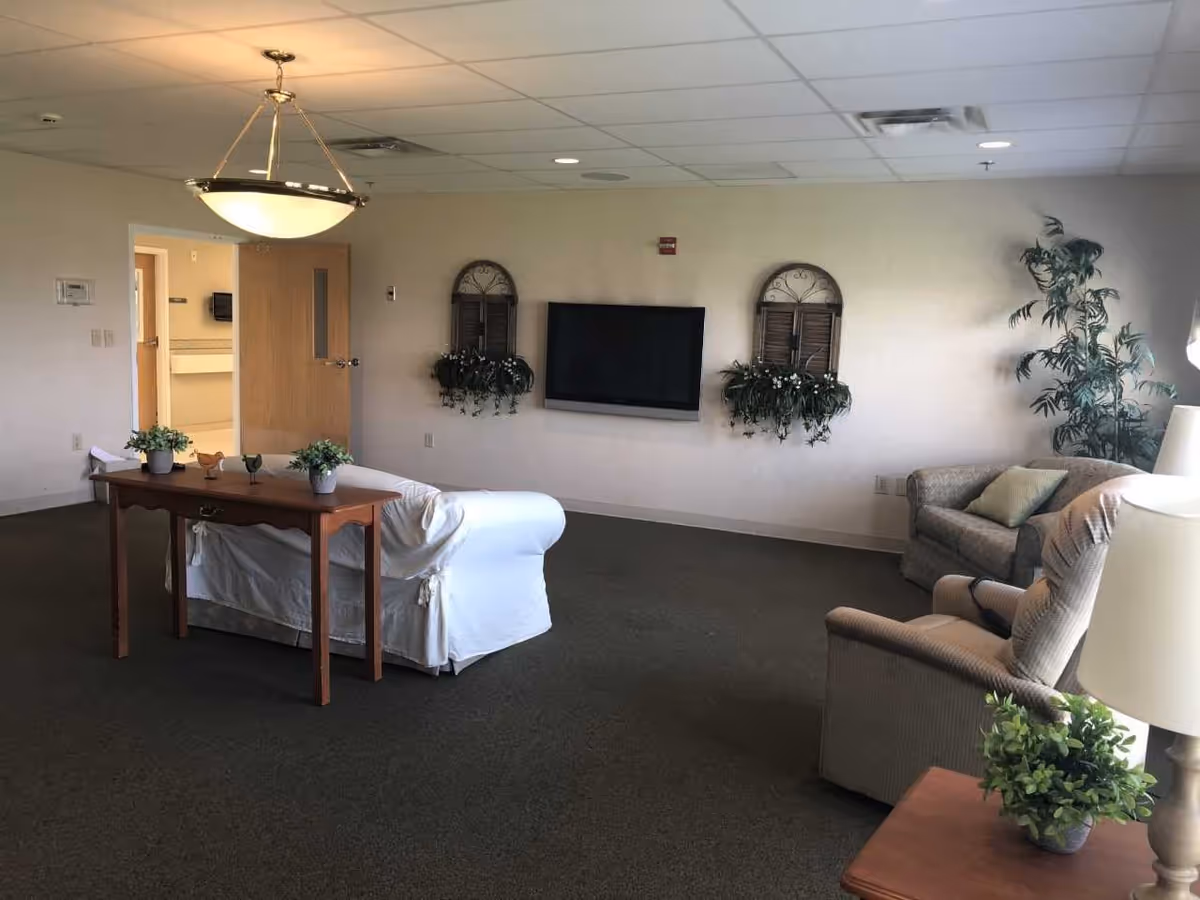 A cozy living room area in a nursing and rehab facility with a white covered sofa, a wooden table with small potted plants and decorative birds, a beige armchair with a pillow, a flat-screen TV mounted on the wall, two wall-mounted decorative plant holders, a tall potted plant in the corner, and a ceiling light fixture. The room has carpeted flooring and neutral-colored walls.
