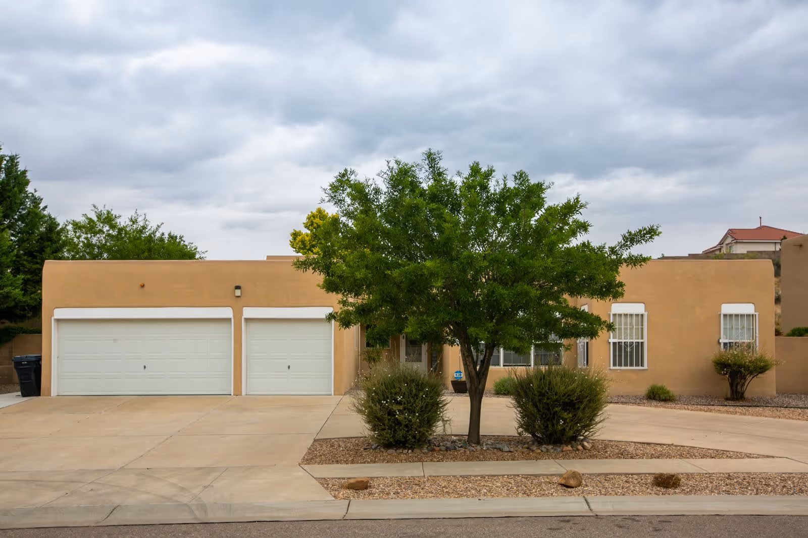 Front exterior view of a single-story beige stucco house with two white garage doors, a tree and bushes in the front yard, and a cloudy sky overhead.