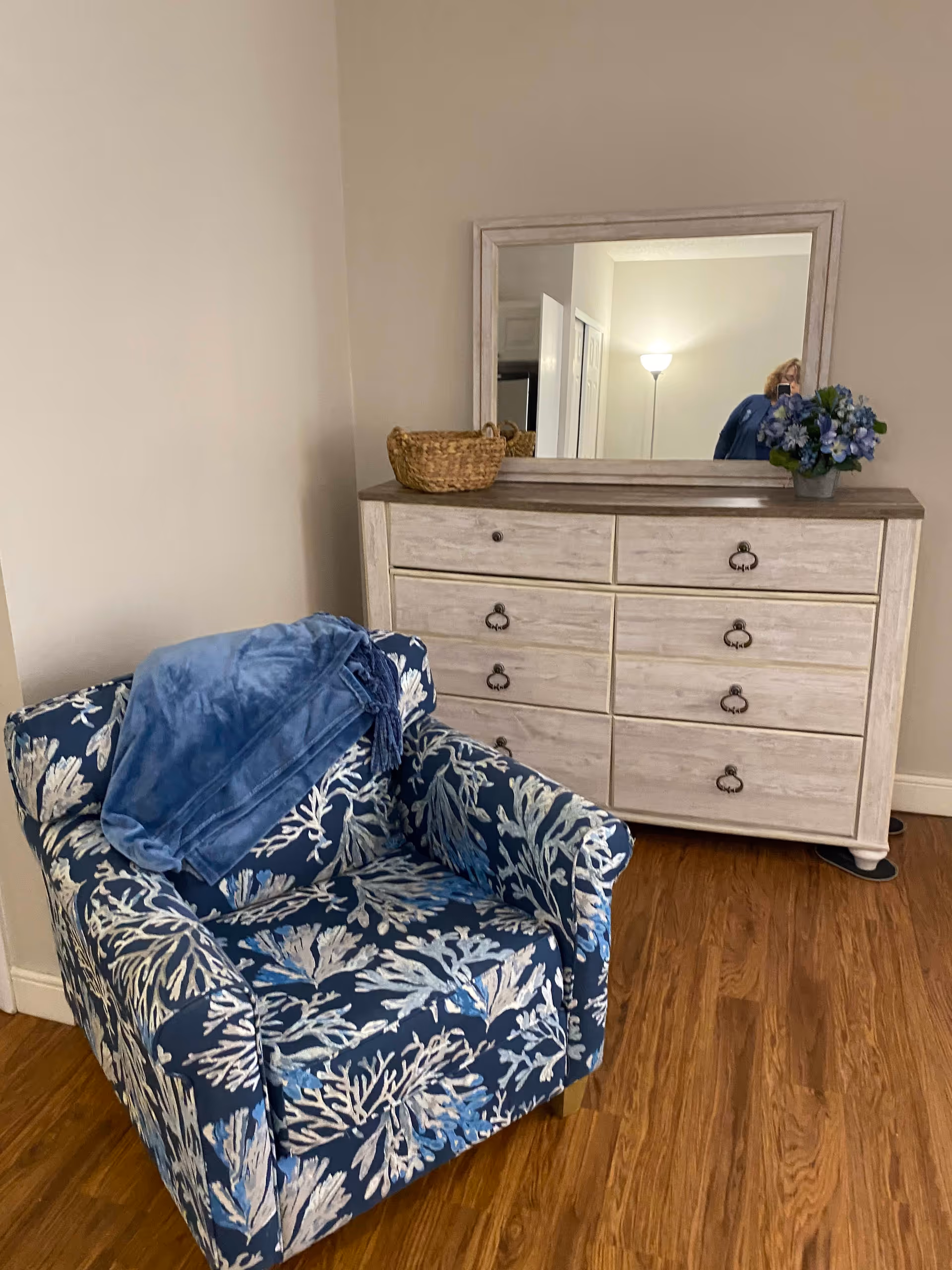 A cozy corner of a room featuring a blue armchair with a white and light blue leaf pattern, draped with a blue blanket. Behind the chair is a light-colored wooden dresser with eight drawers and a large rectangular mirror on top. On the dresser, there is a woven basket and a small pot of blue and purple flowers. The floor is wooden, and a floor lamp and part of a door are visible in the mirror's reflection.