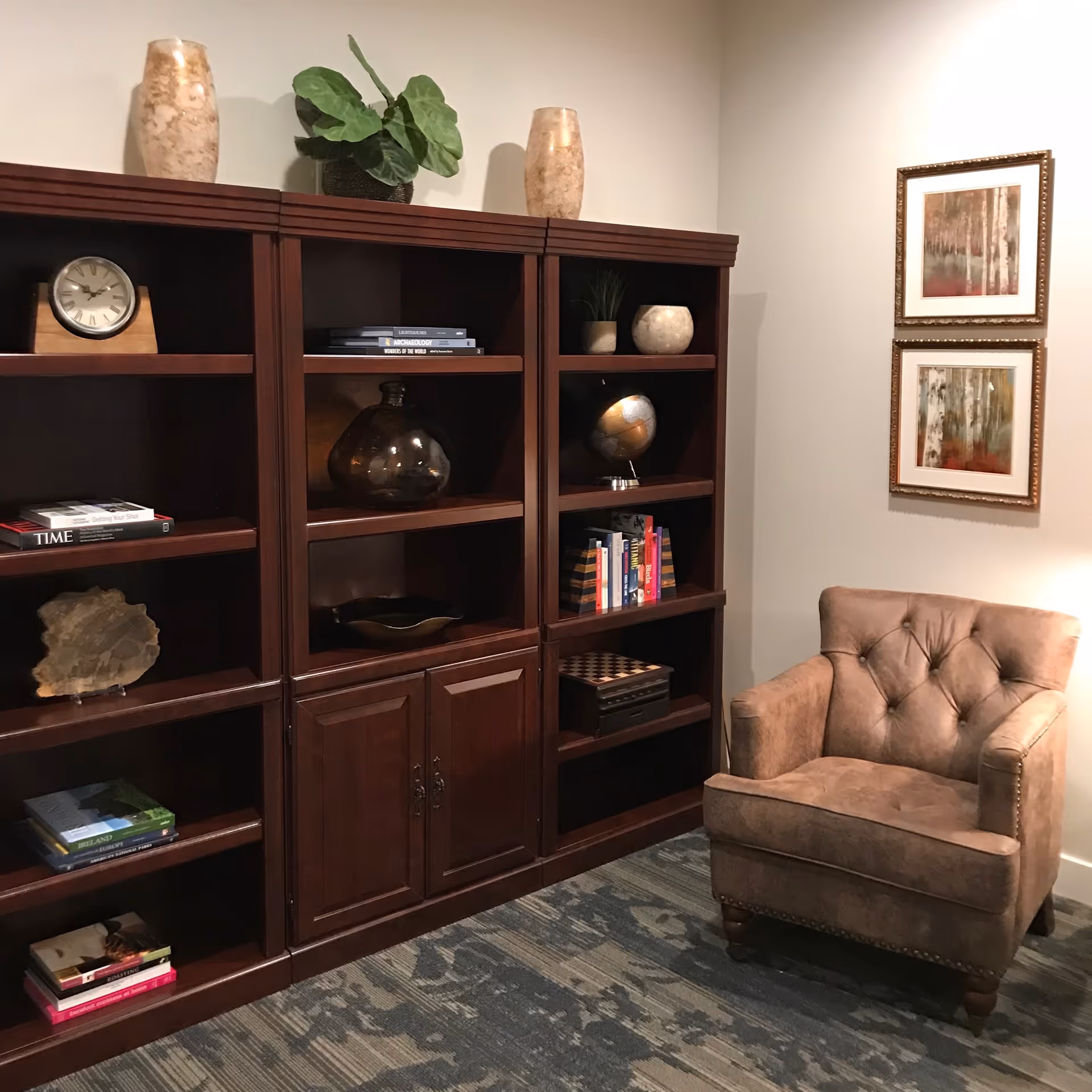 A cozy corner of a room featuring a brown upholstered armchair next to a large wooden bookshelf filled with books, decorative items, a clock, and a globe. Two framed abstract paintings hang on the wall above the chair, and a patterned carpet covers the floor.