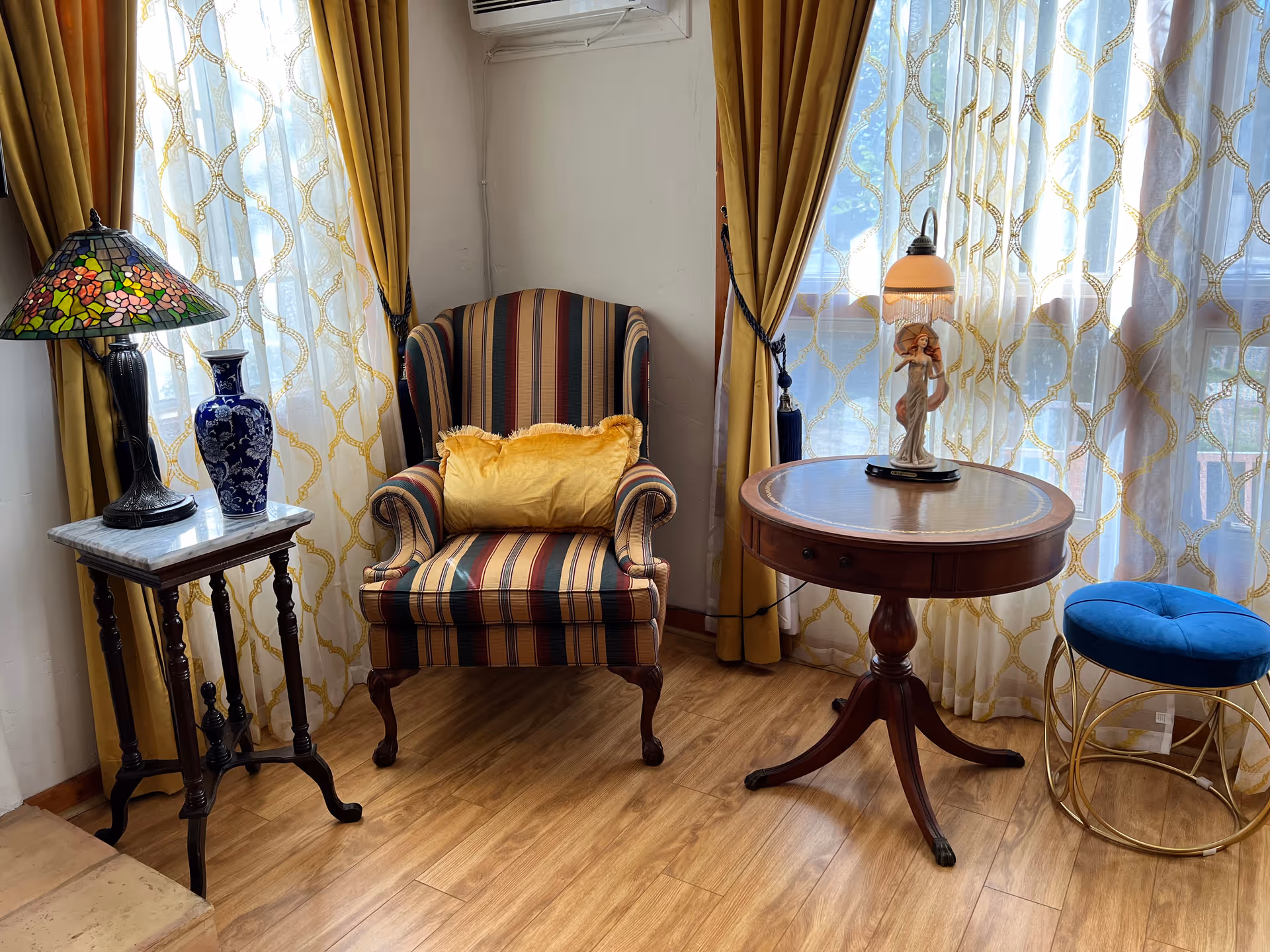 A cozy corner of a living room with a striped armchair featuring a yellow cushion, a round wooden side table with a decorative lamp, a small blue cushioned stool with a gold metal frame, and a small wooden table holding a blue vase and a stained glass lamp. The background shows large windows with sheer white curtains and yellow drapes.