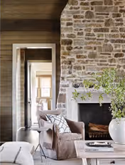 Cozy living room with a stone fireplace, an armchair and coffee table in neutral tones, with a view into an adjoining room.
