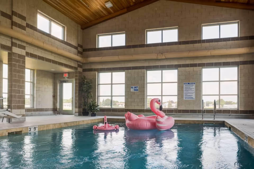 Indoor swimming pool with two pink flamingo floaties in the water. The pool area has large windows letting in natural light, a high wooden ceiling, and beige brick walls. There are pool rules and capacity signs on the wall, and a door leading outside.