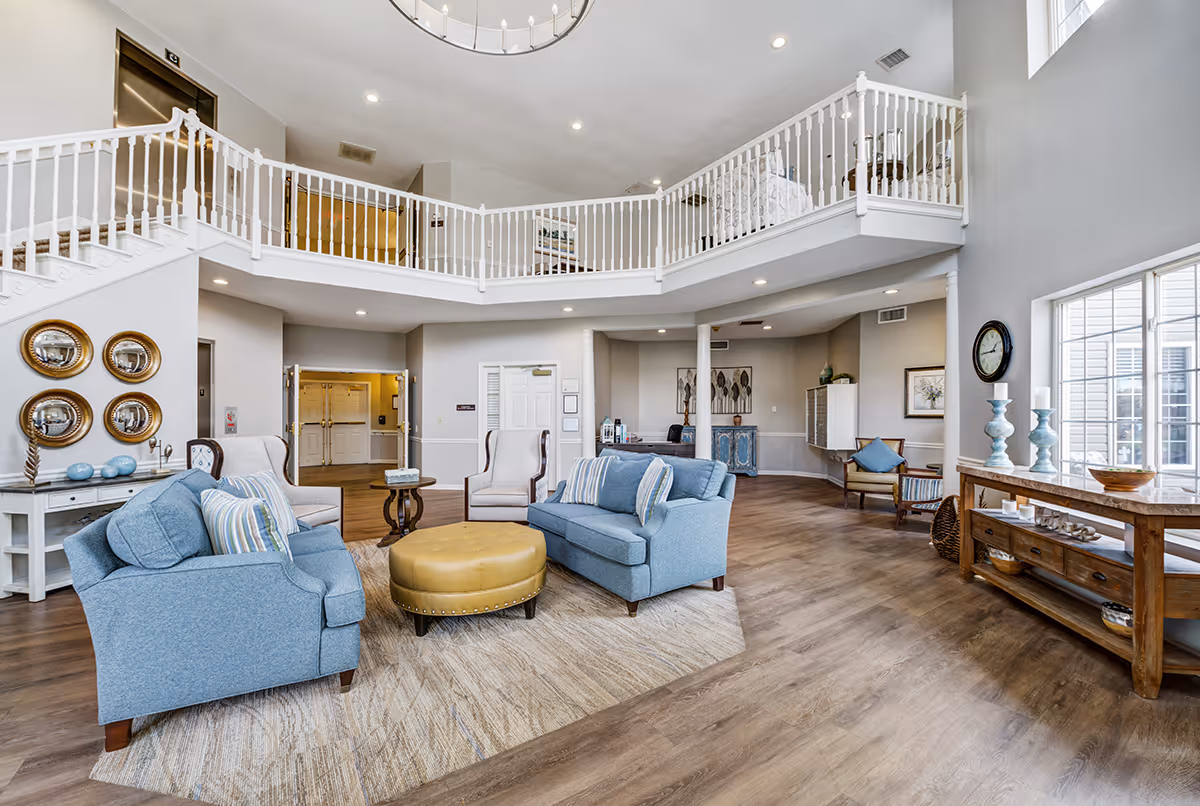 Spacious and bright senior living facility common area with high ceilings, a second-floor balcony railing, two blue sofas, two armchairs, a round yellow ottoman, and various decorative items including wall mirrors, vases, and framed artwork.