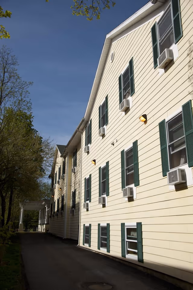 Exterior side view of a multi-story building with cream-colored siding and green shutters on the windows. Several window air conditioning units are installed. The building is adjacent to a paved pathway with trees and greenery on the left side under a clear blue sky.