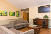 A cozy living room area with two patterned sofas facing a round wooden coffee table. On the wall above a wooden cabinet is a flat-screen TV, and the cabinet holds some electronic devices and a small plant. Three colorful nature-themed paintings hang on the adjacent wall. The room has warm lighting and a wooden floor.