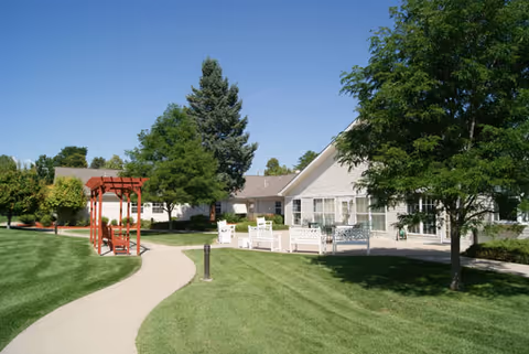 A well-maintained outdoor area at Brookdale Pinehurst Park featuring a curved concrete walkway, green grass, trees, a red wooden pergola with benches, and a white building with large windows under a clear blue sky.
