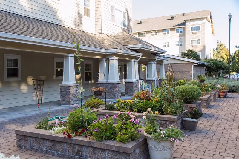 Outdoor garden area at Garden Way Retirement Community featuring raised flower beds with various colorful flowers and plants, a paved walkway, and a building with a covered porch supported by columns in the background.
