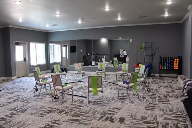 A spacious exercise room with gray walls and patterned carpet, featuring a circle of chairs each equipped with resistance bands and small weights. The room has large windows letting in natural light, a wall mirror, and various exercise equipment including colorful balls and foam rollers in the corner.