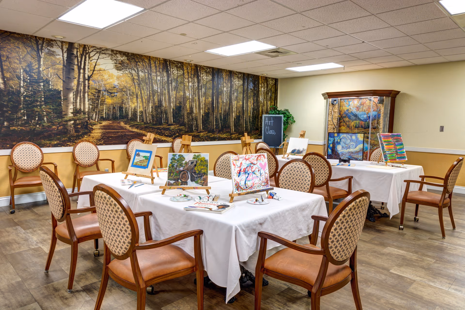 A well-lit room set up for an art class with tables covered in white tablecloths, several chairs with patterned upholstery, and small easels displaying colorful paintings. The room features a large mural of a forest path on one wall and a cabinet with more artwork on display. A small chalkboard sign reads 'Art Class'.