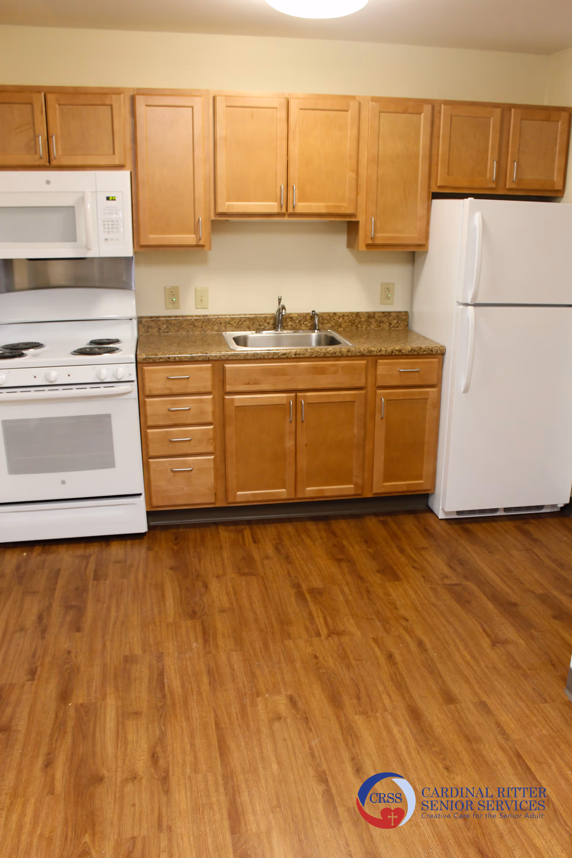 A kitchen area with wooden cabinets, a white stove with an oven, a white microwave above the stove, a stainless steel sink with a faucet, and a white refrigerator. The floor is wood-patterned, and the walls are light-colored.