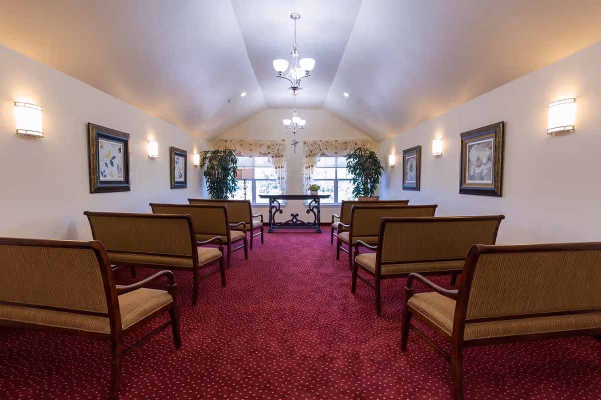 Symmetrical chapel-like interior with rows of wooden benches on red carpet facing a windowed alcove and chandelier lighting.
