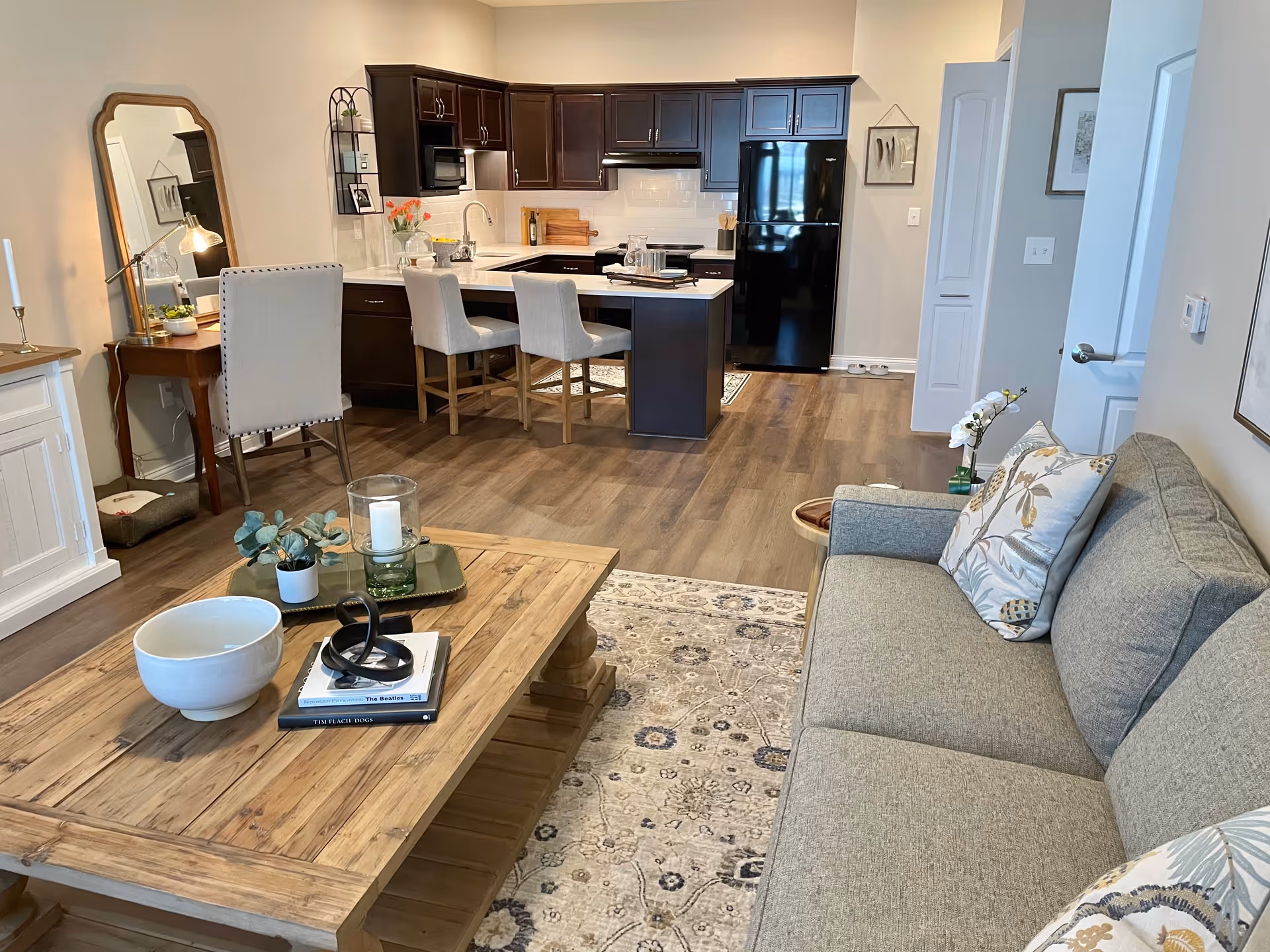 A modern open-concept living area featuring a gray sofa with patterned pillows, a wooden coffee table with decorative items, and a kitchen in the background with dark cabinets, a black refrigerator, and a white countertop with three upholstered bar stools. The room has wood flooring and neutral-colored walls with framed artwork.