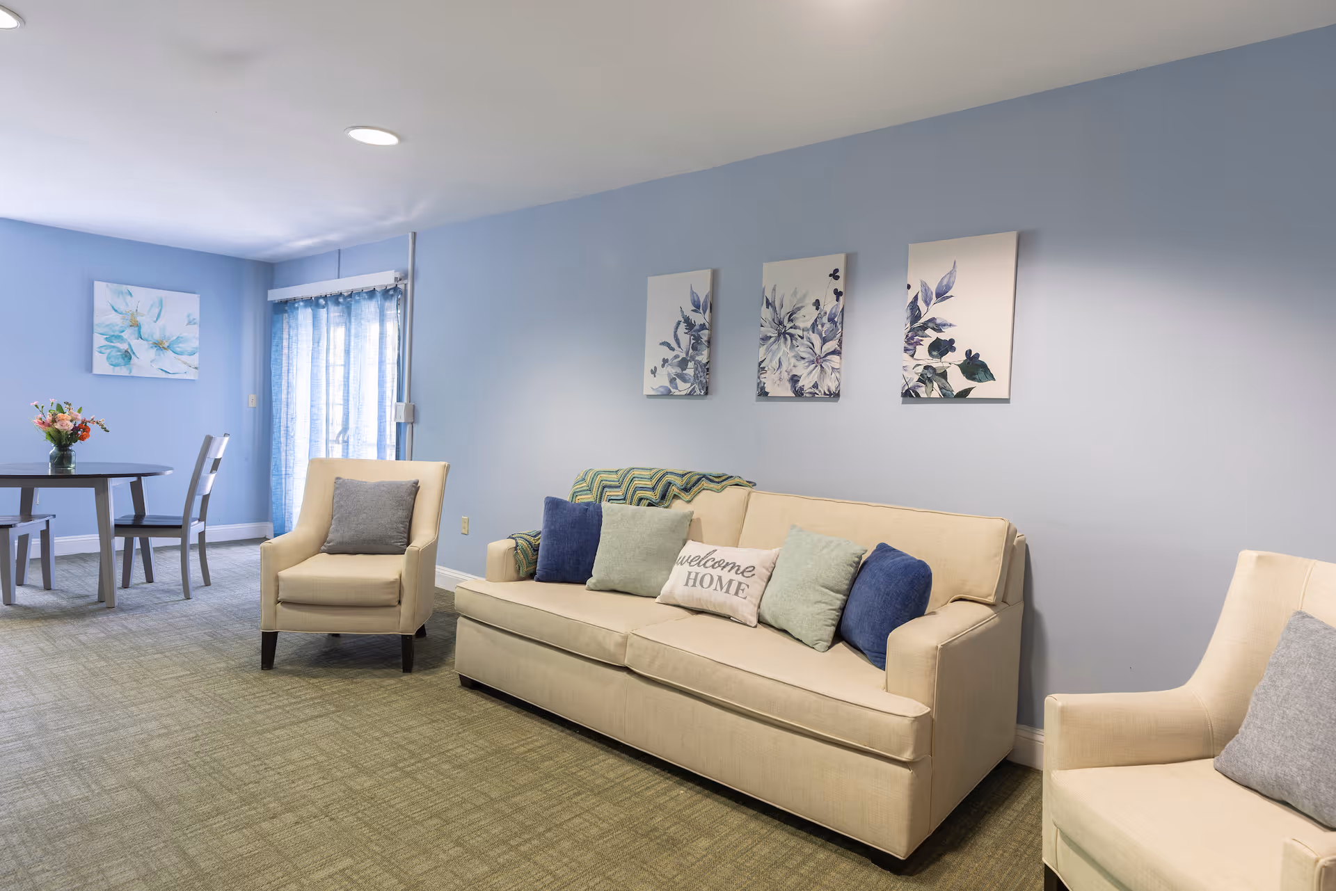 A cozy living room with light blue walls, featuring a beige sofa adorned with multiple cushions including one that says 'welcome HOME'. There are two beige armchairs with gray cushions, a round dining table with chairs in the background, and three floral paintings on the wall above the sofa. A window with sheer blue curtains allows natural light into the room.