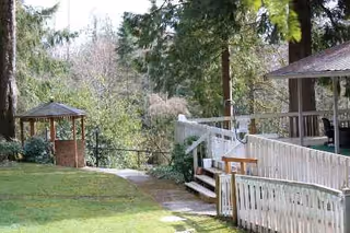 Outdoor view of a walkway with white railing, gazebo, and grassy area among trees.