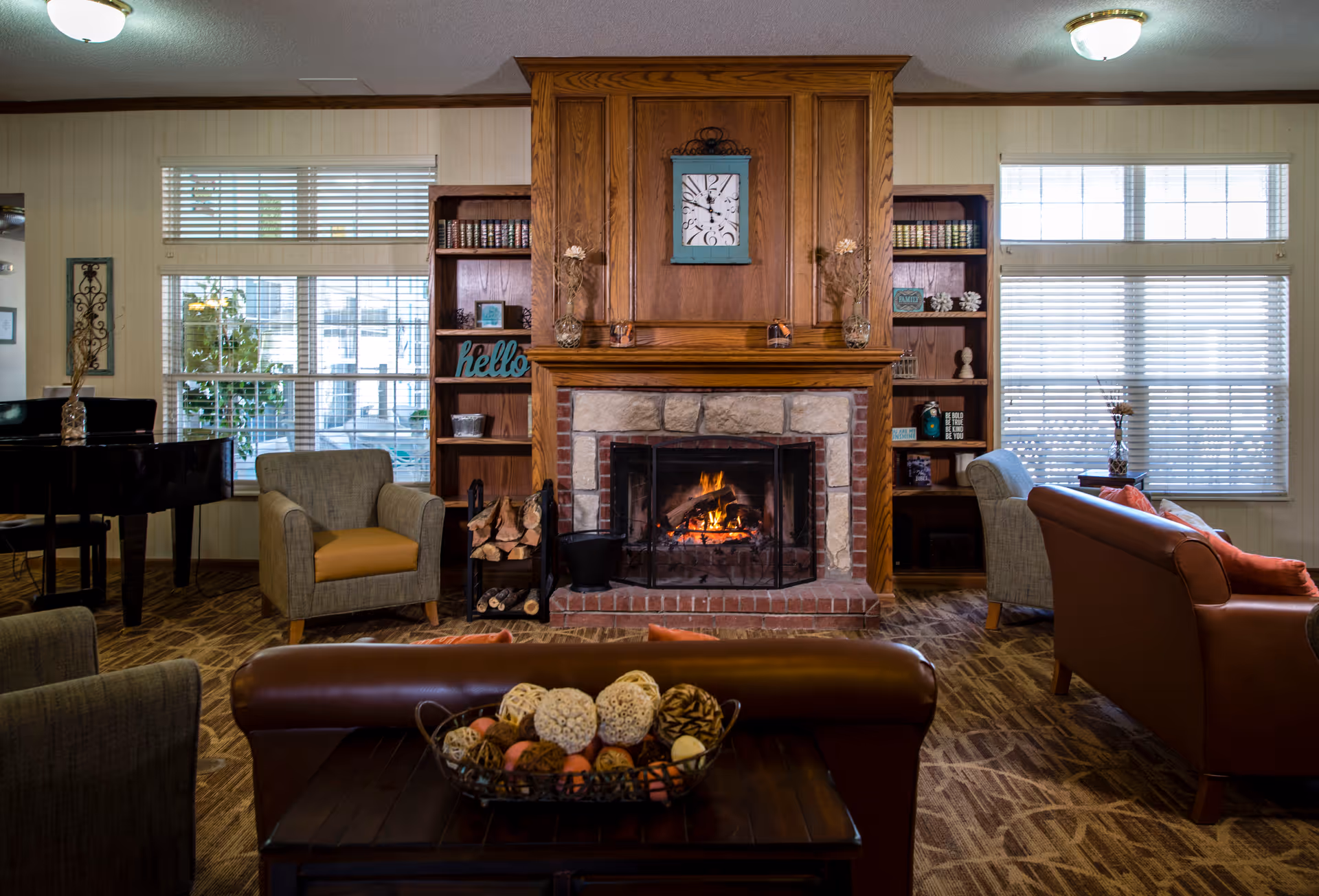 Cozy living room with a central wood-paneled fireplace, seating, bookshelves, and large windows.