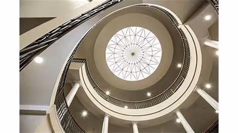 View looking upward inside a building showing a circular spiral staircase with white columns and a large round skylight with a geometric pattern at the top.