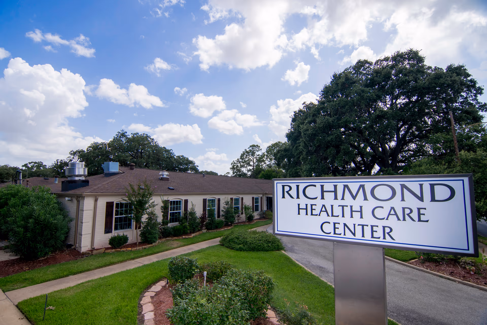 Exterior view of Richmond Health Care Center building with a large sign in the foreground displaying the facility's name. The building is single-story with multiple windows and surrounded by green landscaping and trees under a partly cloudy sky.