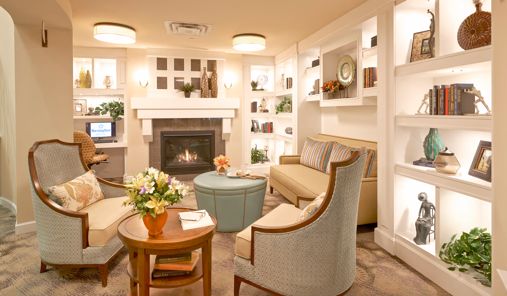 A cozy living room area with a fireplace in the center, flanked by built-in shelves filled with decorative items and books. The room features a beige sofa with striped pillows, two upholstered armchairs with floral cushions, a round wooden table with a flower vase, and a blue ottoman. Soft lighting creates a warm and inviting atmosphere.