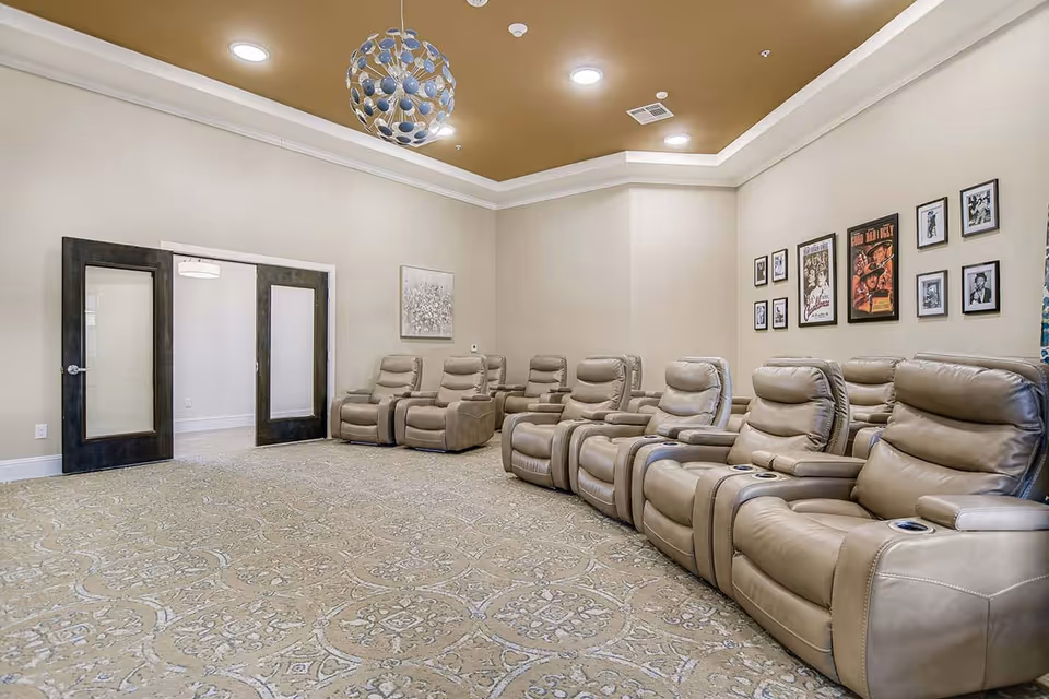 A room with two rows of beige leather recliner chairs arranged facing the same direction, likely for watching movies or presentations. The room has a patterned carpet, beige walls with framed pictures and movie posters, a decorative ceiling light fixture, and double glass doors.