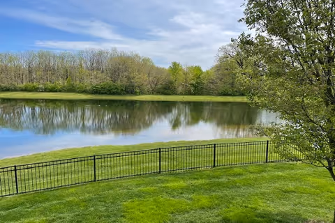 A peaceful outdoor scene featuring a calm pond surrounded by green grass and trees under a partly cloudy sky. A black metal fence runs along the edge of the grassy area near the water.