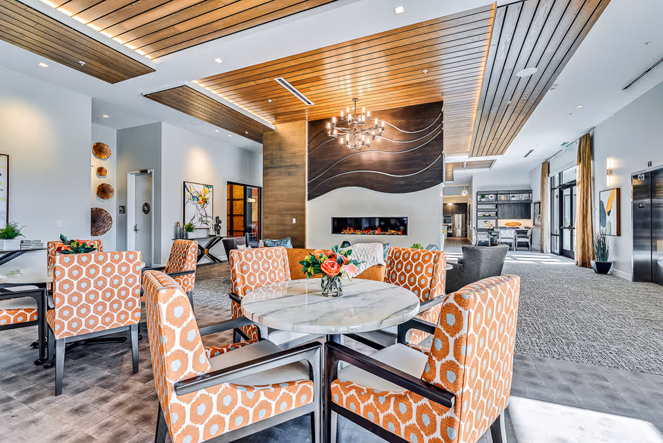 Bright, modern assisted living common area with patterned orange chairs around a round marble table, a fireplace, and lounge seating under wood-paneled ceiling accents.