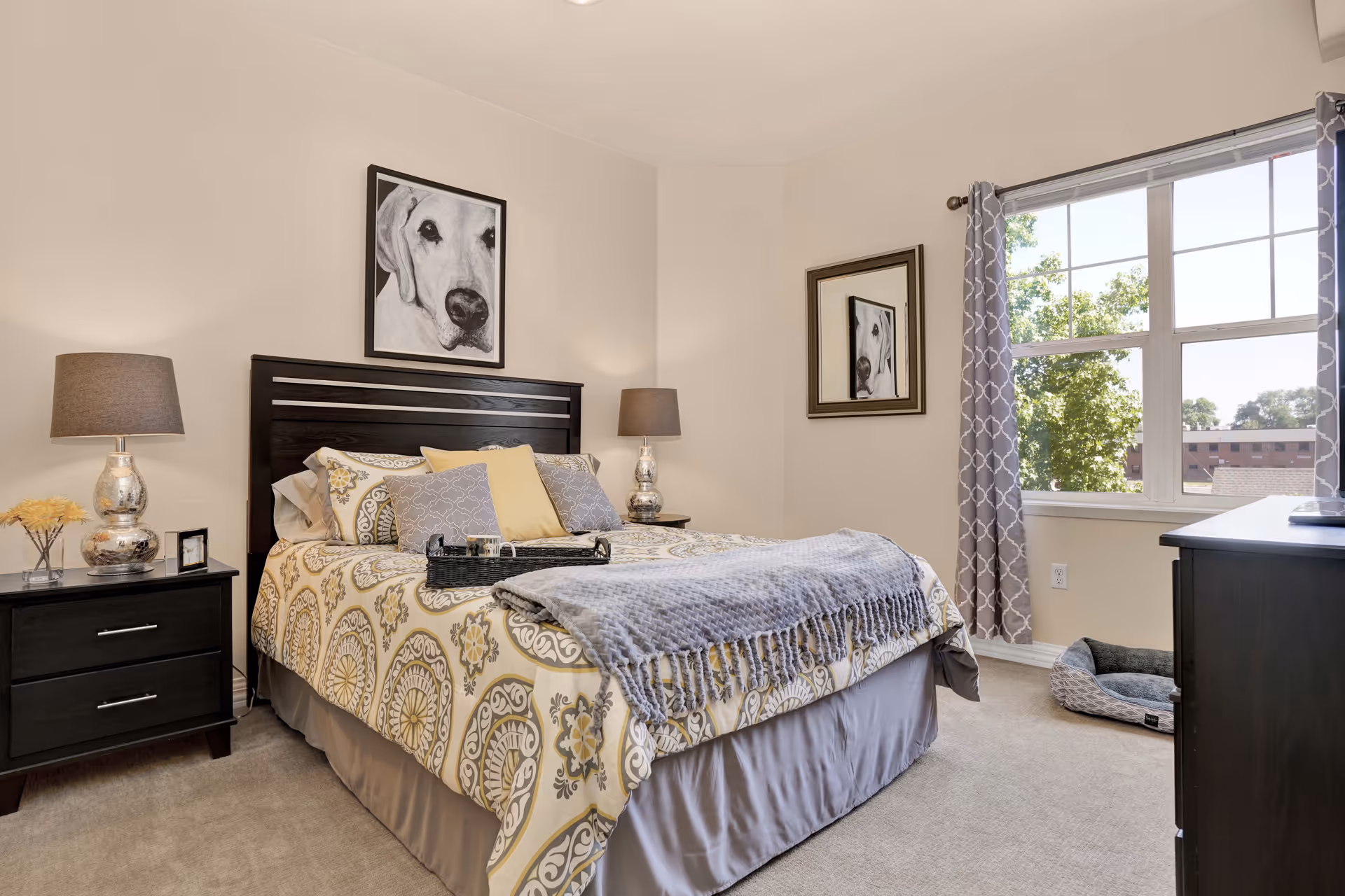 A well-lit bedroom with a large bed featuring patterned yellow and gray bedding, multiple pillows, and a gray throw blanket. There are two matching bedside tables with silver lamps and a small flower arrangement on one. A framed black and white dog portrait hangs above the headboard, and a matching mirror is on the adjacent wall. A window with patterned gray curtains lets in natural light, and a pet bed is placed on the carpeted floor near the window.