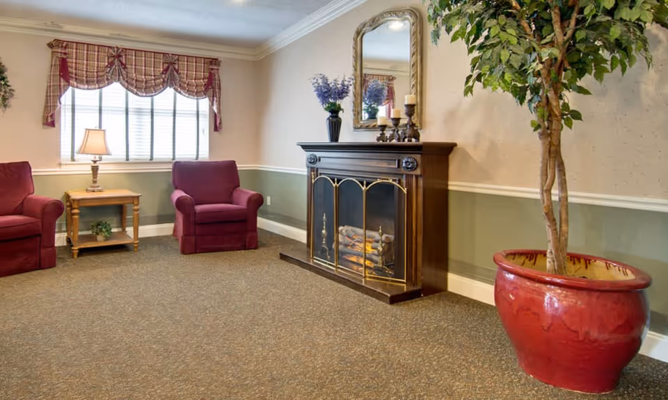 A cozy sitting area in a nursing center featuring two red armchairs, a wooden side table with a lamp, a decorative fireplace with candles and a vase on top, a large mirror above the fireplace, and a large potted plant in a red ceramic pot. The room has a window with plaid valance curtains and beige walls with a green wainscoting.
