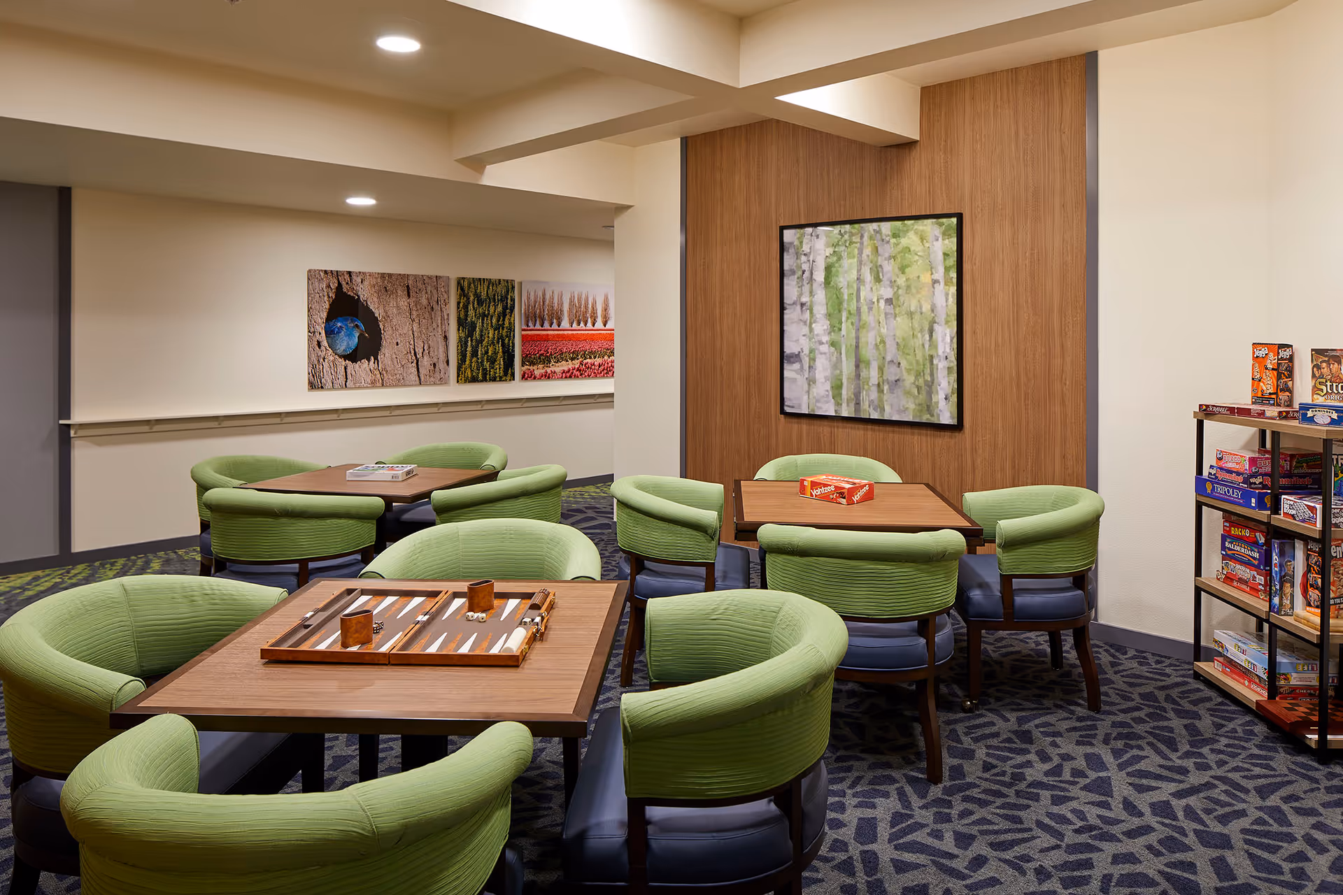 A cozy game room with several square wooden tables surrounded by green cushioned chairs. On the tables are board games including backgammon and Yahtzee. The room has patterned carpet, neutral walls with nature-themed artwork, and a shelf filled with various board games.