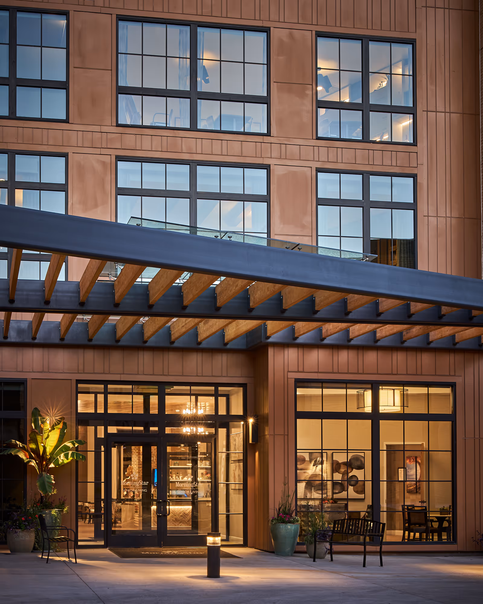 Entrance of MorningStar Senior Living at Old Town building at dusk, featuring large glass doors and windows, a wooden pergola overhead, potted plants, benches, and warm interior lighting visible through the windows.