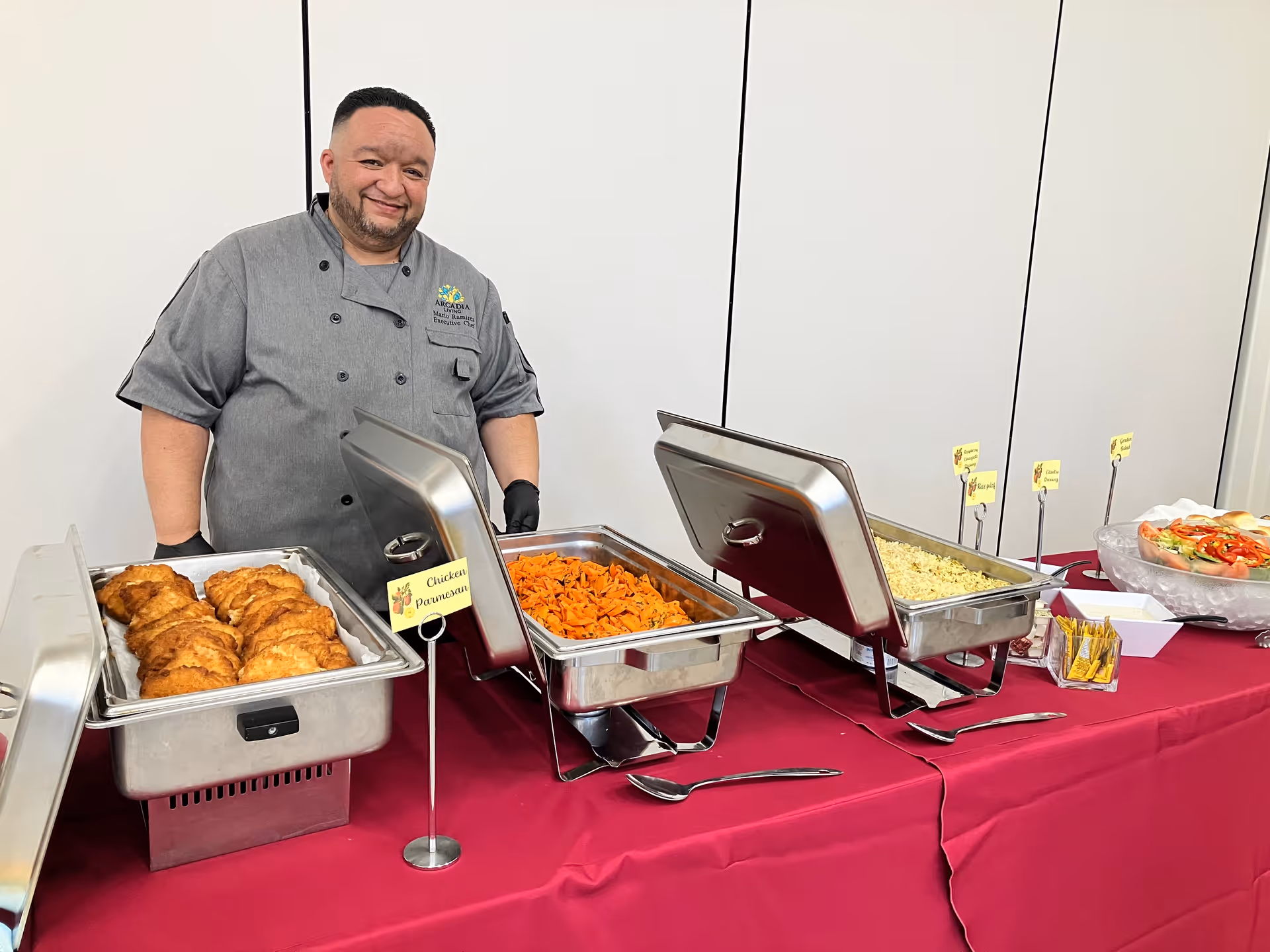 A chef wearing a gray uniform stands behind a buffet table covered with a red tablecloth. The buffet includes trays of chicken parmesan, cooked carrots, rice, and a salad. The chef is smiling and wearing black gloves.