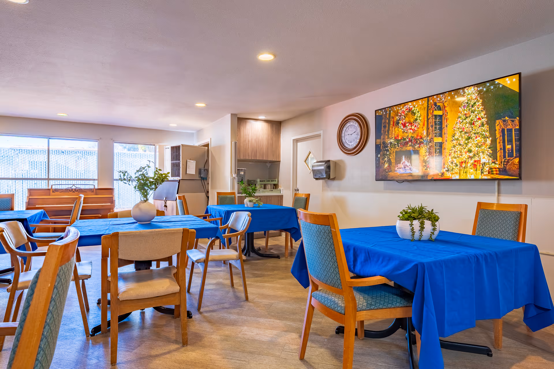 A dining room with several tables covered in blue tablecloths, each surrounded by wooden chairs with cushioned seats. There are small potted plants on the tables. A large flat-screen TV on the wall displays a festive holiday scene with a decorated Christmas tree and a fireplace. A clock and a paper towel dispenser are mounted on the wall. Large windows allow natural light to fill the room.