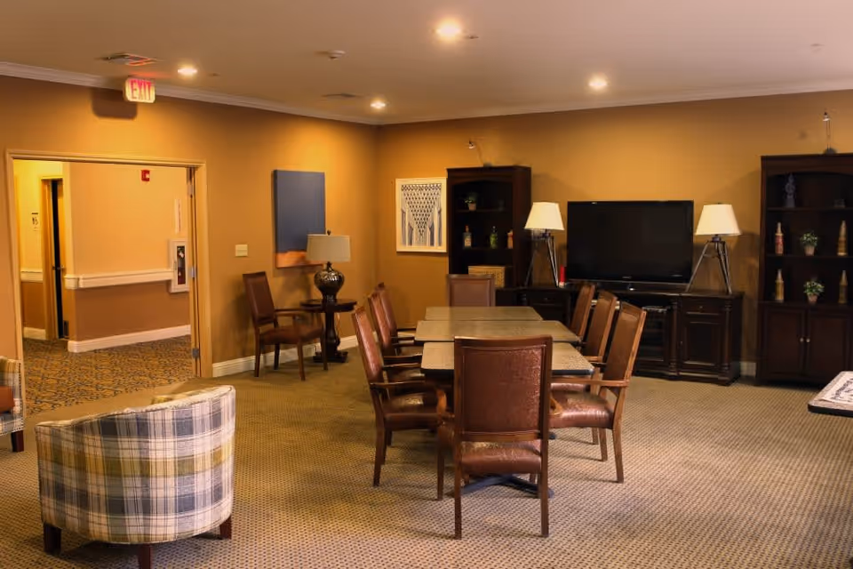 A cozy common area with a rectangular table surrounded by eight brown leather chairs. The room has warm beige walls and carpeted floors. There are two wooden bookshelves flanking a flat-screen TV on a wooden TV stand. Two table lamps with white shades are placed on either side of the TV. A small round table with a lamp and two chairs is positioned near an open doorway leading to a hallway. Two plaid upholstered armchairs are partially visible in the foreground.