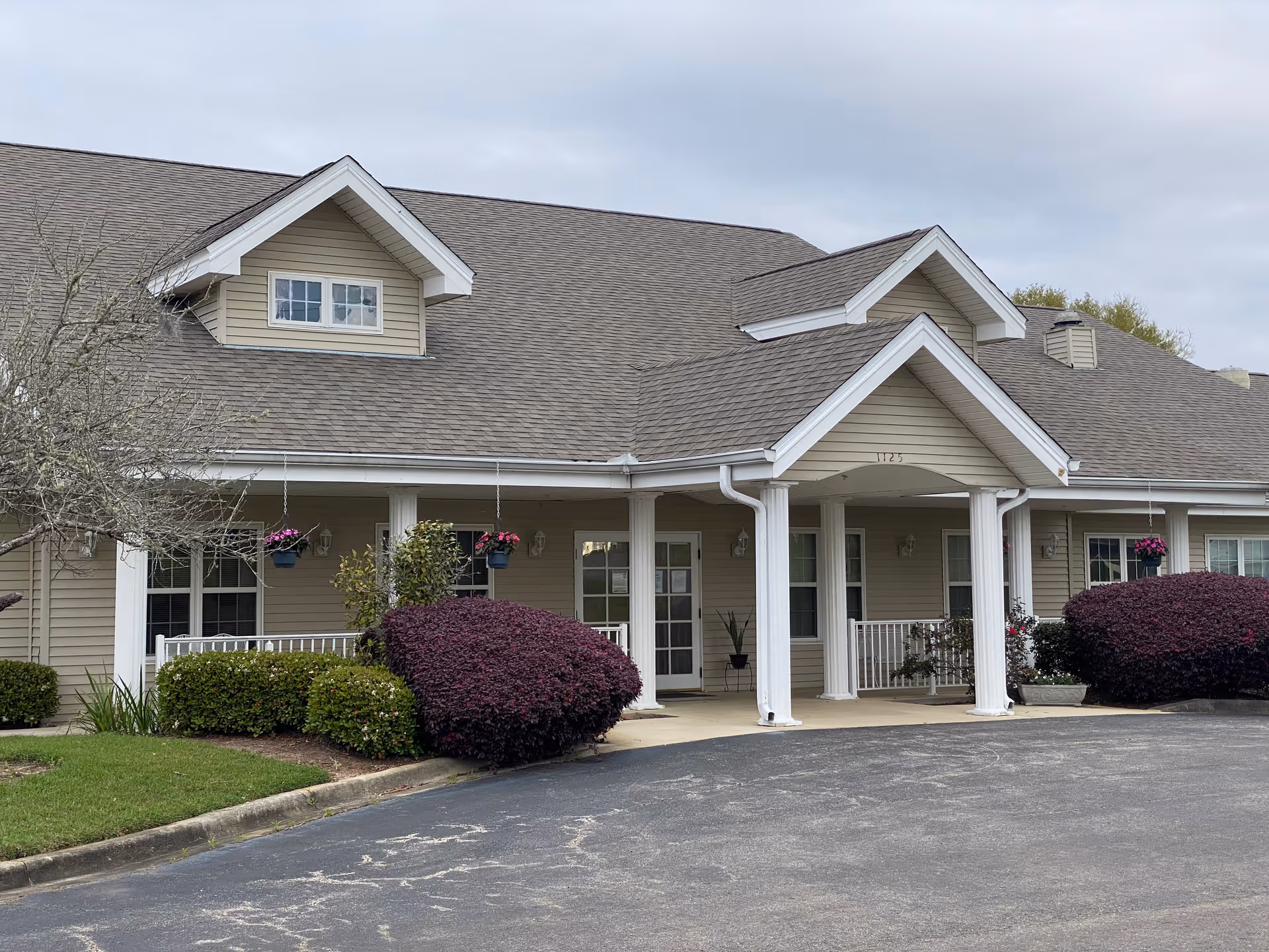 Exterior view of a single-story beige building with a gray shingled roof, white columns, and a covered entrance. There are bushes and hanging flower pots near the entrance, and a paved driveway in front.