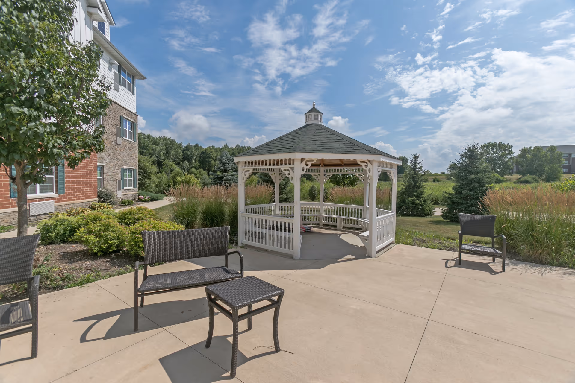 Outdoor patio area at Deer Path of Huntley featuring a white gazebo with decorative trim, surrounded by greenery and trees. There are several black wicker chairs and a small table on the concrete patio under a partly cloudy blue sky.
