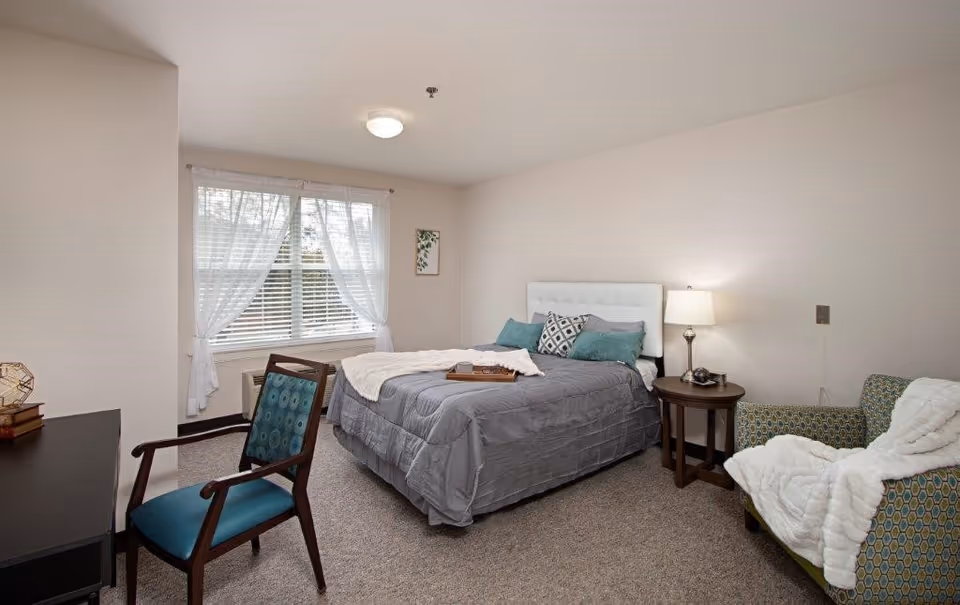 A bright assisted-living bedroom with a bed, bedside table and lamp, chairs, desk, and a window with sheer curtains.