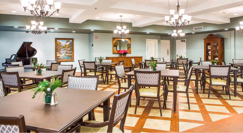 A spacious dining room with multiple tables and chairs arranged neatly. Each table has a small floral centerpiece. The room features chandeliers hanging from the ceiling, a grand piano in the corner, a large painting on the wall, and a wooden cabinet with glass doors displaying dishes. The floor has a patterned design with wood and tile.