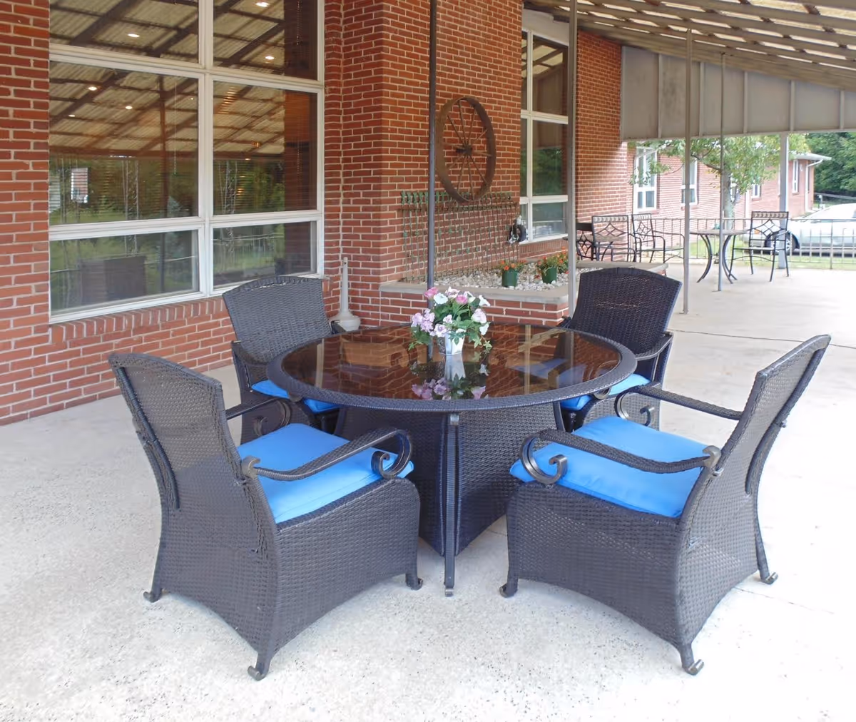 Outdoor covered patio area with a round glass-top table surrounded by four black wicker chairs with blue cushions. The patio has a brick wall with large windows and decorative elements, including a wagon wheel mounted on the wall and small potted plants. Additional tables and chairs are visible in the background.