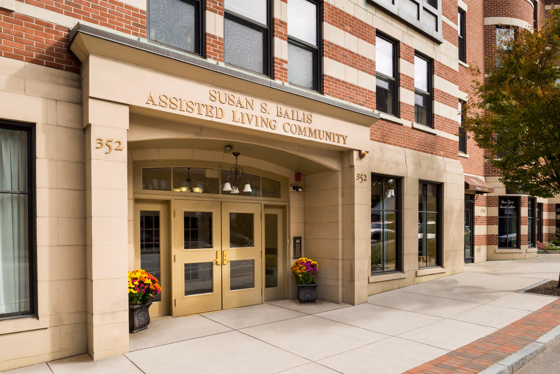Entrance of the Susan S. Bailis Assisted Living Community showing gold double doors, the building name above, address number 352, and potted flowers.