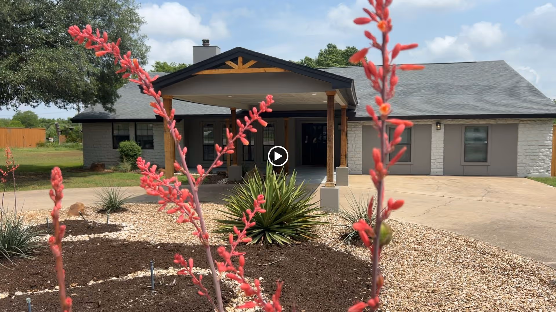 Front exterior view of a single-story senior living facility building with a covered entrance supported by wooden pillars. The foreground features a landscaped area with red flowering plants, green shrubs, and gravel. The sky is partly cloudy.