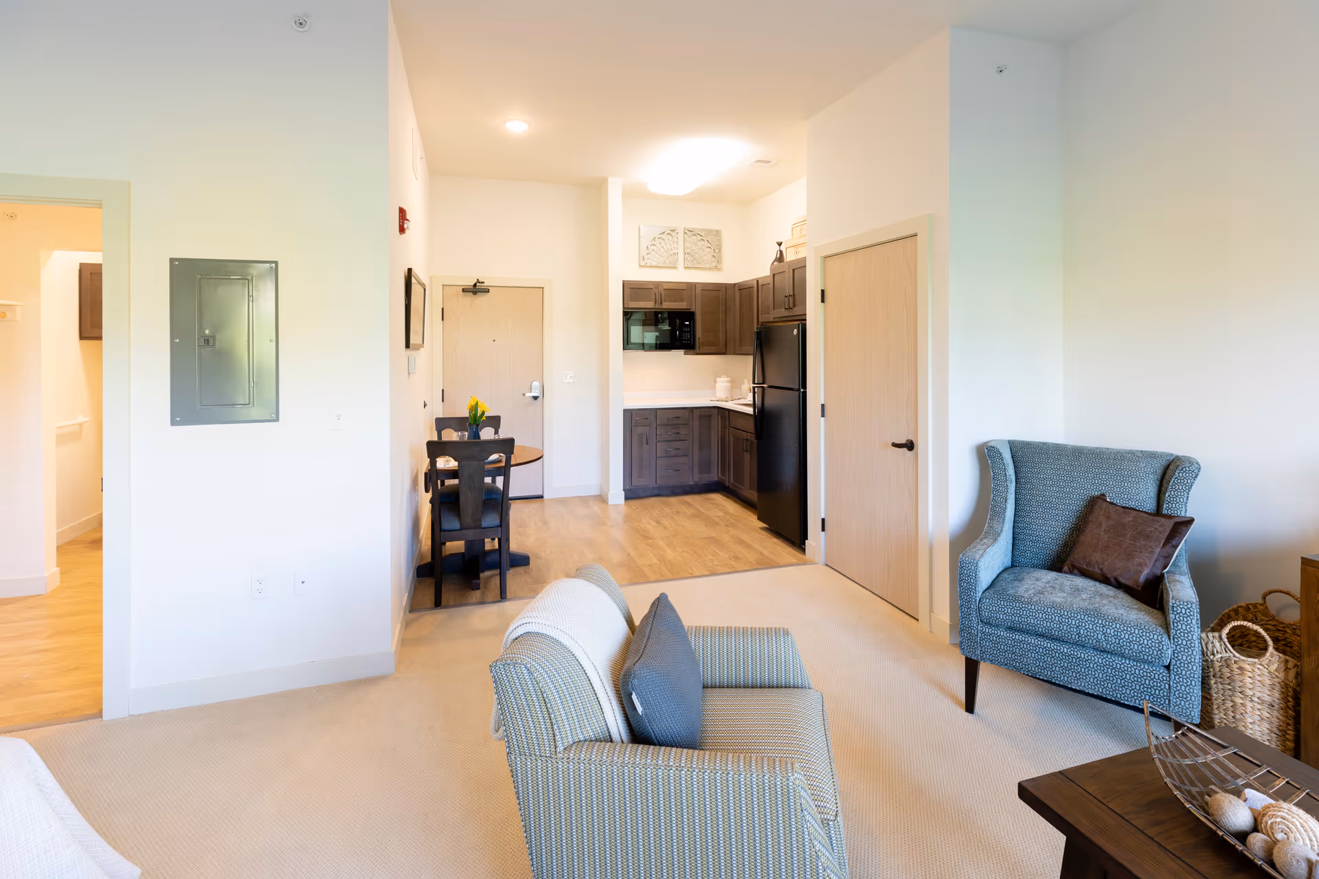 A bright and clean senior living apartment interior featuring a small kitchen with dark cabinets, a black refrigerator, and a microwave. Adjacent to the kitchen is a small dining area with a round wooden table and four chairs. The living area includes a patterned armchair with a brown pillow, a loveseat with cushions, and a wooden coffee table with decorative items. Light-colored walls and carpeted flooring complete the cozy space.