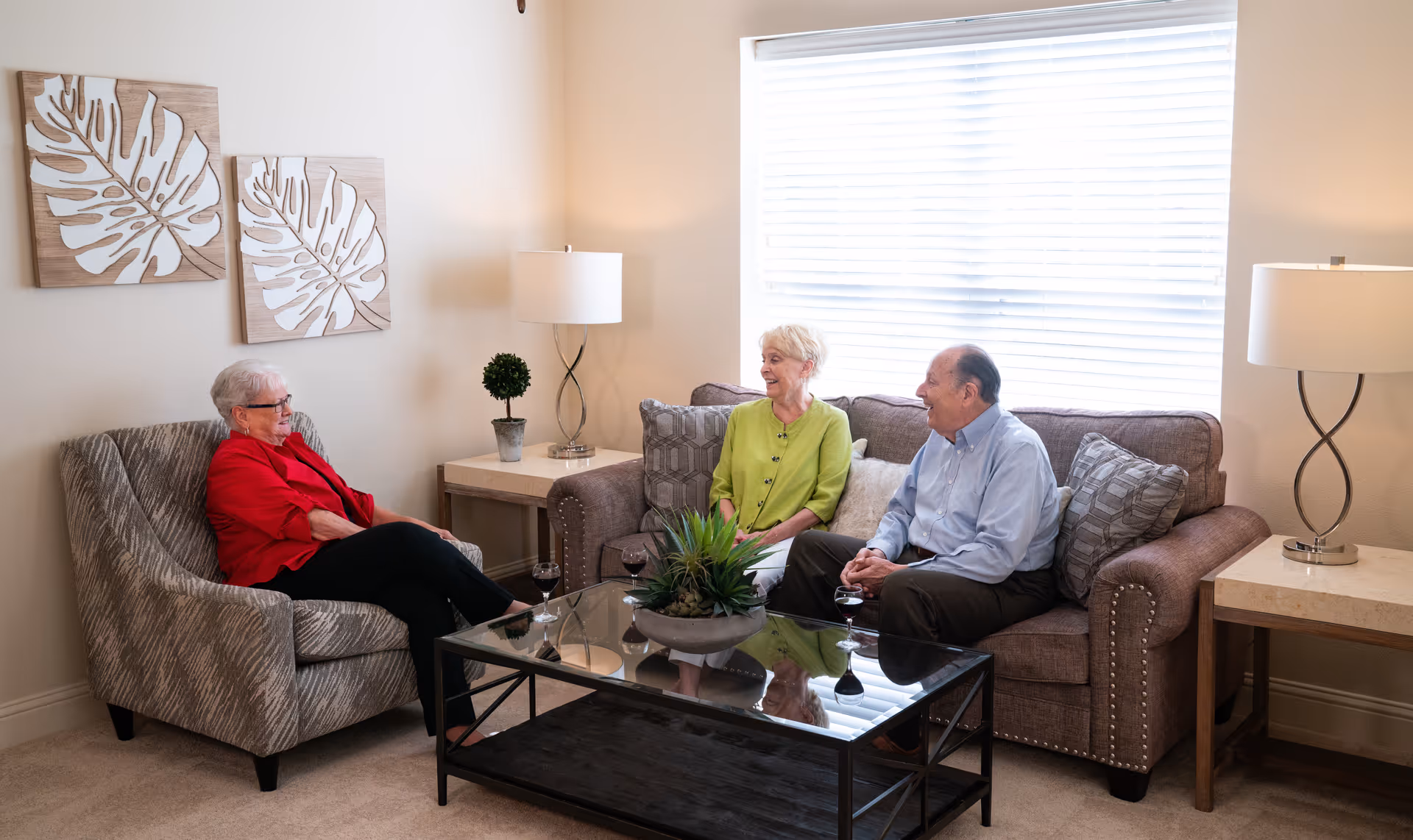 Three elderly people sitting and chatting in a well-lit living room. Two are seated on a brown sofa with patterned cushions, and one is seated on a gray patterned armchair. There is a glass coffee table with a plant centerpiece and three glasses of red wine. The room has beige walls, two decorative leaf wall art pieces, two side tables with modern lamps, and a large window with blinds.